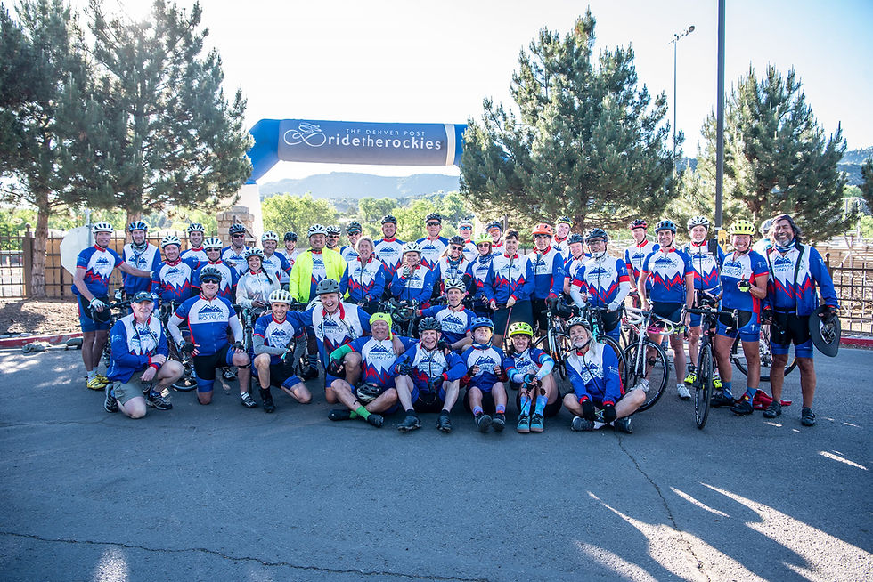 Cyclists in matching blue and white jerseys pose under a "Ride the Rockies" banner, smiling against a backdrop of trees and mountains.