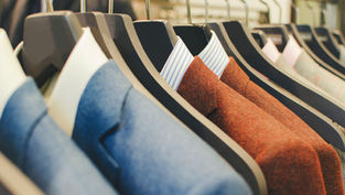 Row of suits on hangers, featuring blue and red jackets with striped shirts. Displayed in a retail setting, creating a formal, elegant vibe.