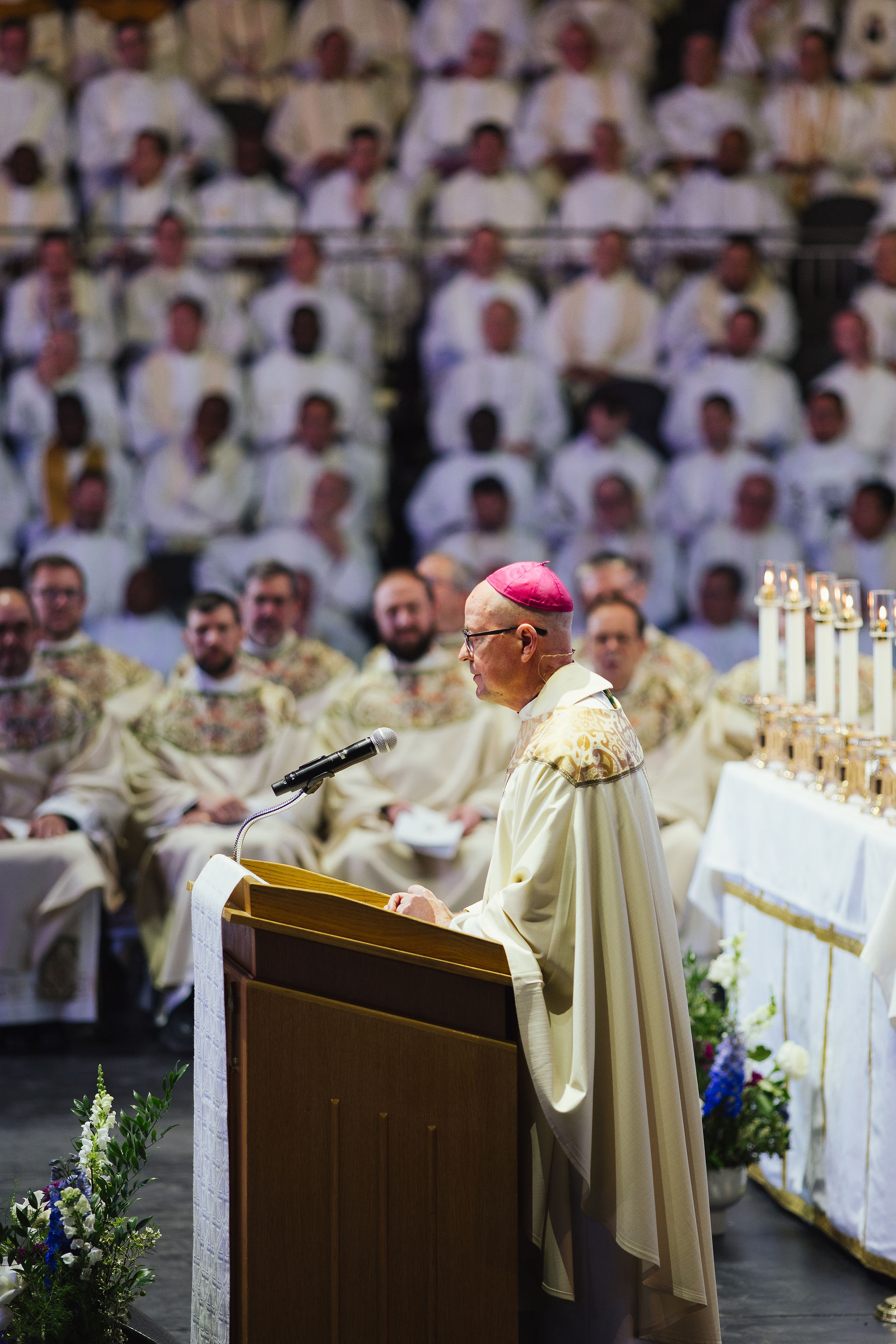 A bishop in ornate robes speaks at a podium. A crowd of clergy in white robes is seated in the background. Candles and flowers adorn the altar.