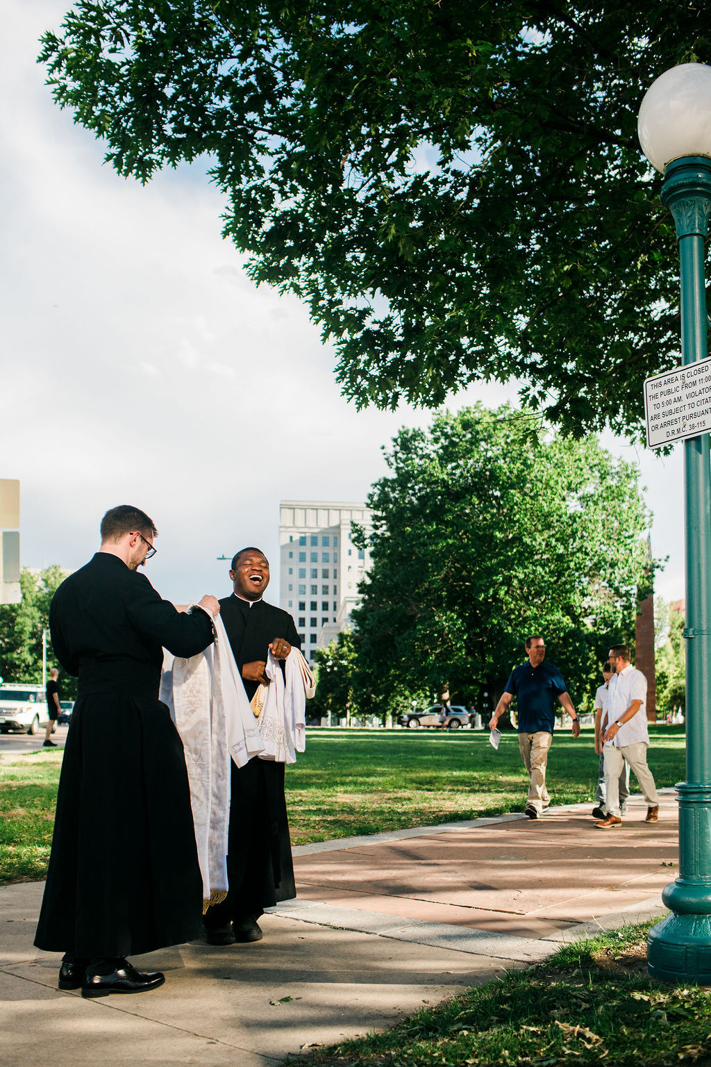 Two priests in black robes laugh on a tree-lined park path, with people walking by and a posted closed-area sign on a green lamppost.