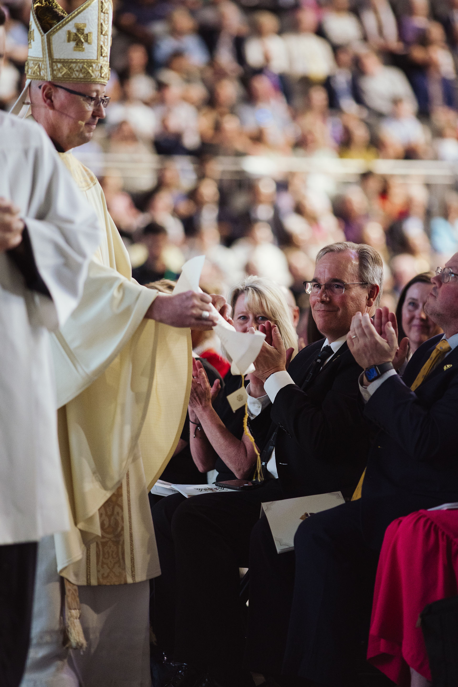 A bishop in gold vestments hands a document to a seated attendee as people around them applaud during a liturgical ceremony.
