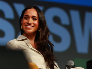Smiling woman with long brown hair wearing a light shirt, on stage with "SXSW" text in background. Warm lighting, microphone visible.