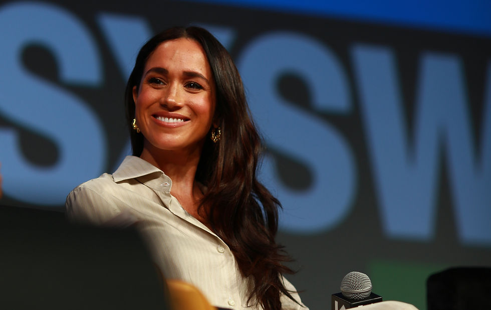 Smiling woman with long brown hair wearing a light shirt, on stage with "SXSW" text in background. Warm lighting, microphone visible.