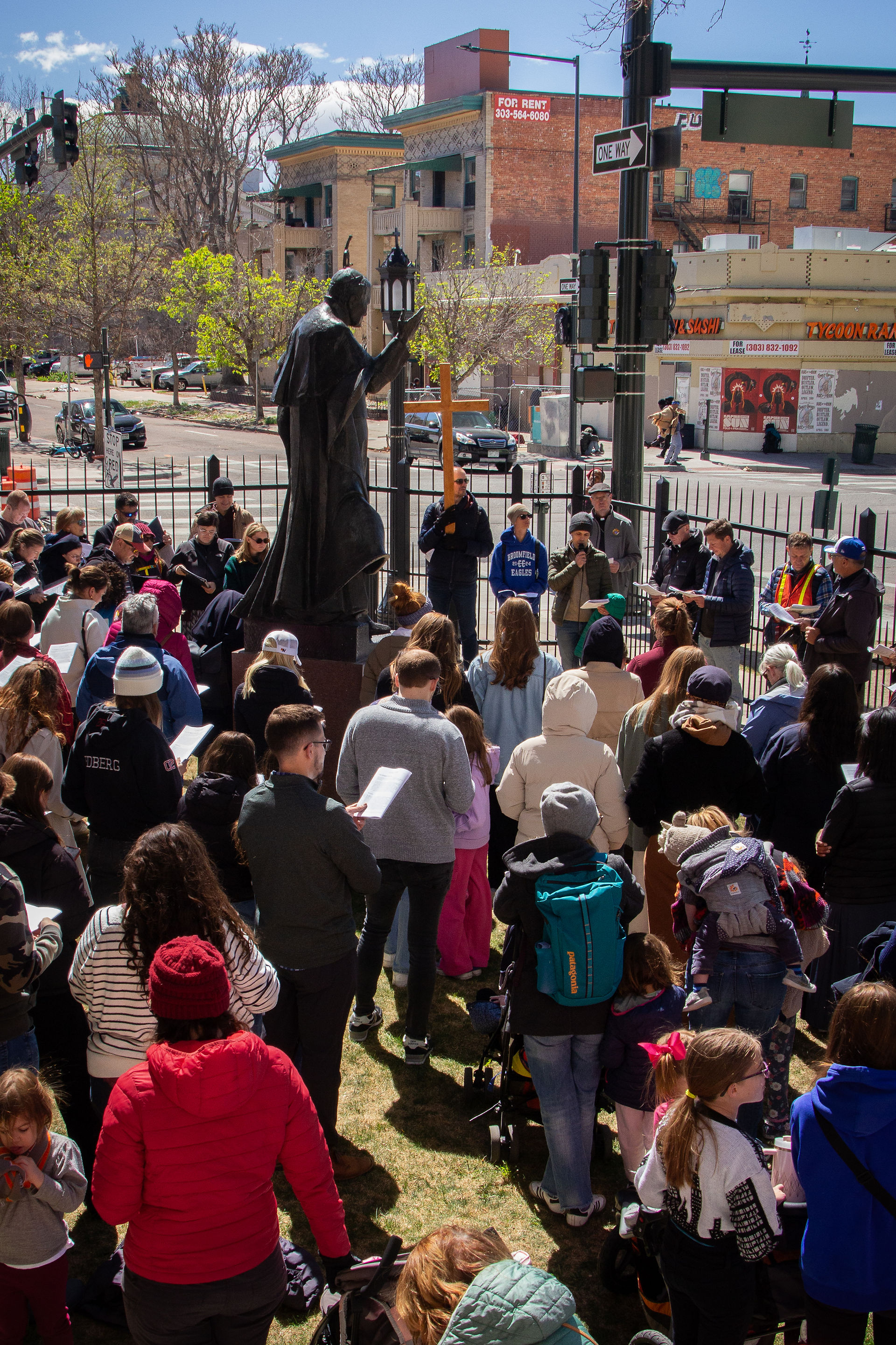 A group gathers on church steps around a wooden cross, holding papers and praying together outdoors.