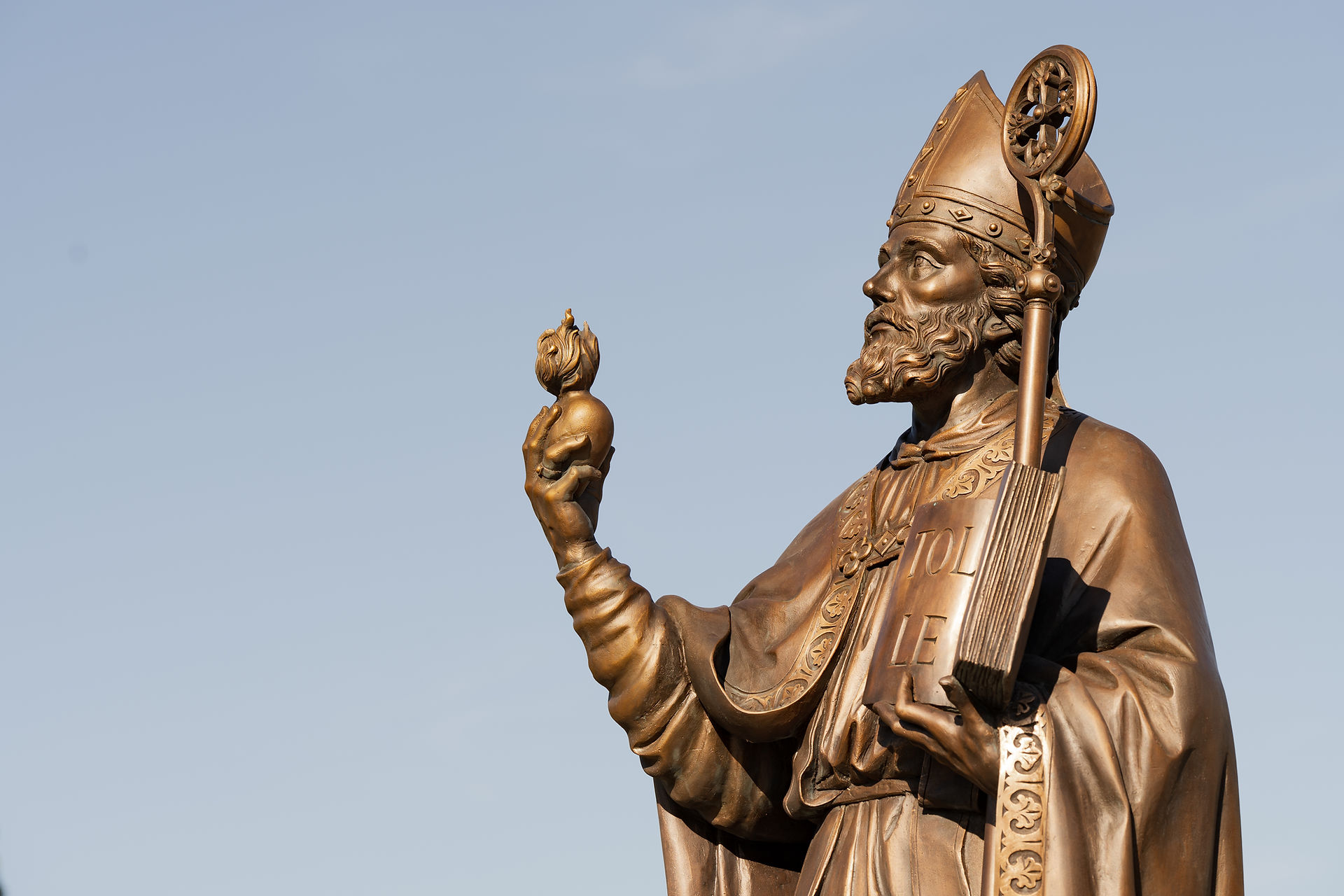 Bronze statue of a bishop holding a heart and a book with "Tolle" text, set against a clear blue sky. He looks determined and wise.