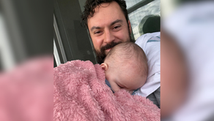 Smiling man cradles sleeping baby wrapped in a pink fuzzy blanket on a porch by a window, a tender, cozy moment.
