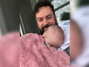 Smiling man cradles sleeping baby wrapped in a pink fuzzy blanket on a porch by a window, a tender, cozy moment.
