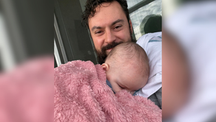 Smiling man cradles sleeping baby wrapped in a pink fuzzy blanket on a porch by a window, a tender, cozy moment.