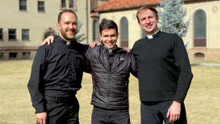 Three seminarians smiling, arm in arm, outside a historic building with arched windows. Wearing black attire; sunny day, relaxed mood.