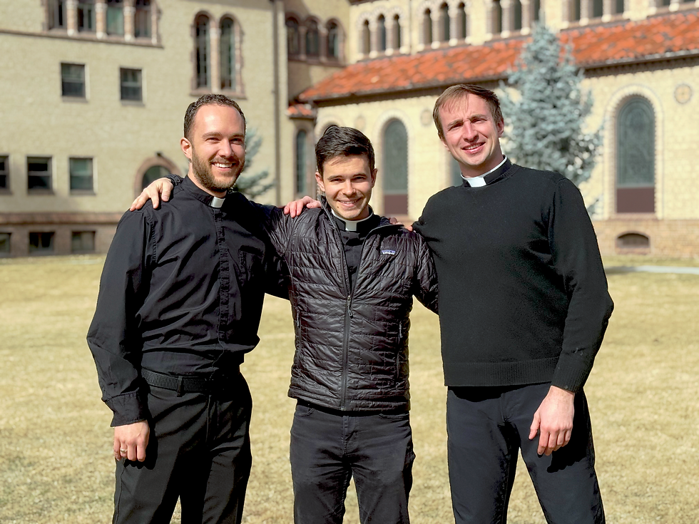 Three seminarians smiling, arm in arm, outside a historic building with arched windows. Wearing black attire; sunny day, relaxed mood.