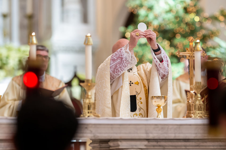 Bishop in ornate robes holds up Eucharist in a church with candles and gold chalices. Soft lighting and bokeh effect in background.