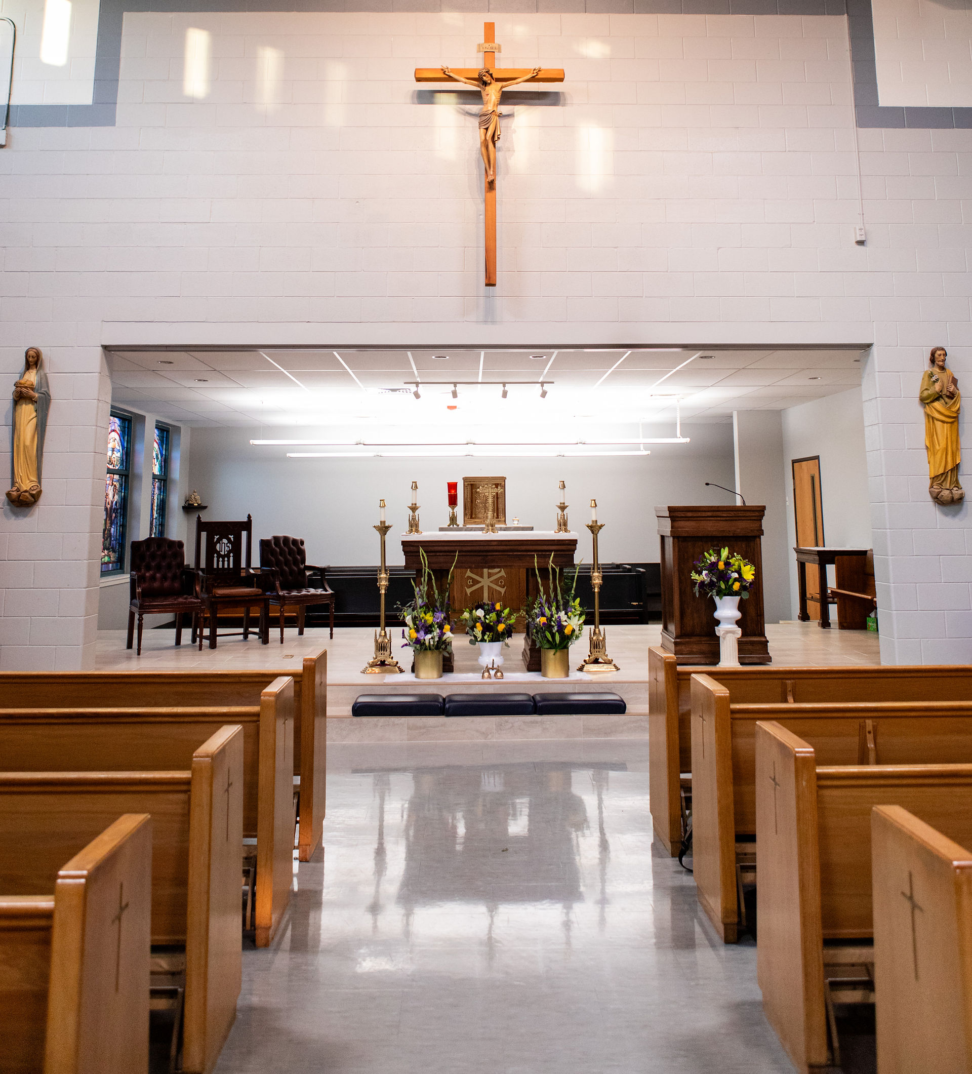 Church interior with wooden pews, an altar adorned with flowers, and a crucifix on the wall. Statues flank the altar. Calm, reverent atmosphere.