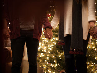 People holding hands in front of a lit Christmas tree. Cozy attire with a warm atmosphere; sweaters and scarves are visible.