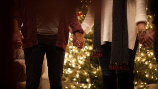 People holding hands in front of a lit Christmas tree. Cozy attire with a warm atmosphere; sweaters and scarves are visible.