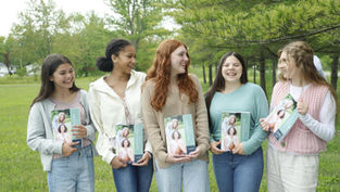 Five smiling women holding books titled "Friendships" stand outdoors on grass with trees in the background, all wearing casual outfits.