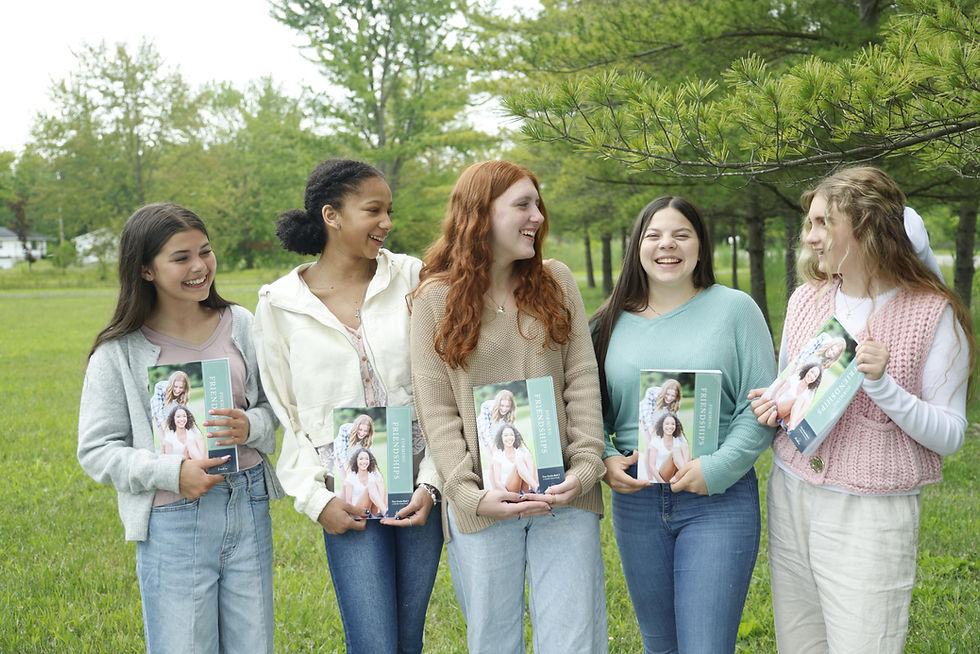 Five smiling women holding books titled "Friendships" stand outdoors on grass with trees in the background, all wearing casual outfits.