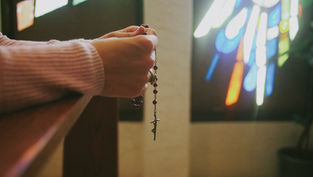 Hands holding a rosary in a dimly lit room with colorful stained glass windows. Peaceful mood, soft light creating a serene atmosphere.