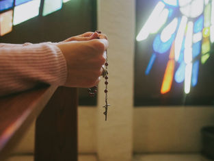 Hands holding a rosary in a dimly lit room with colorful stained glass windows. Peaceful mood, soft light creating a serene atmosphere.