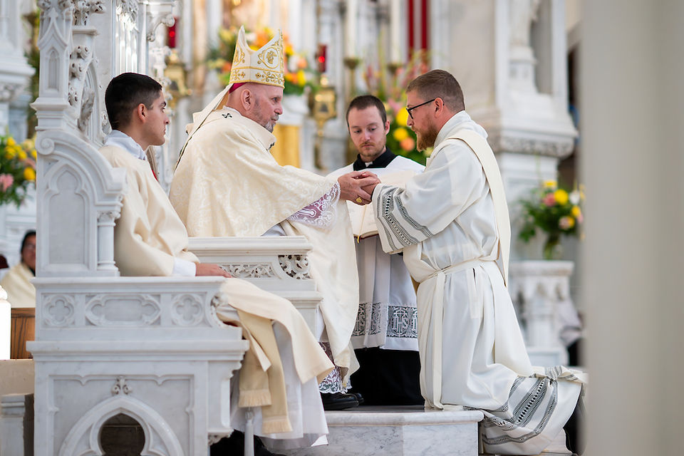 A bishop in ornate robes and hat blesses a kneeling man in white amid a floral-decorated church, with two others observing solemnly.