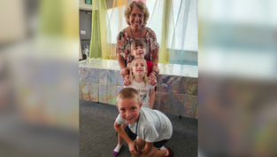 Smiling woman with three children posing indoors under pastel drapes; EXIT sign and shiny silver backdrop behind them.