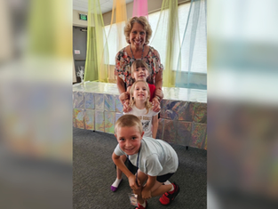 Smiling woman with three children posing indoors under pastel drapes; EXIT sign and shiny silver backdrop behind them.
