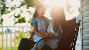 Mother and child sit on a porch, bathed in warm sunlight. The girl wears blue overalls, both smiling at each other, surrounded by greenery.