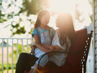 Mother and child sit on a porch, bathed in warm sunlight. The girl wears blue overalls, both smiling at each other, surrounded by greenery.