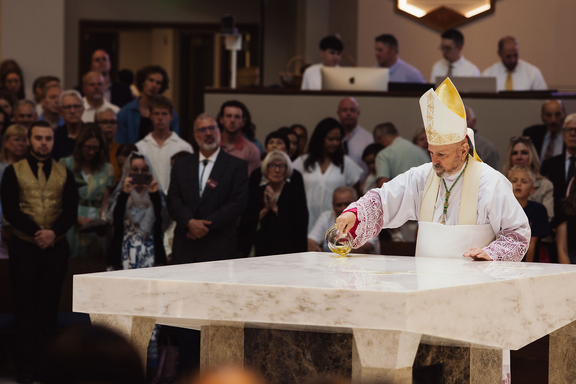 A bishop anoints a marble altar with oil in a church ceremony. A crowd watches solemnly in the softly lit background.