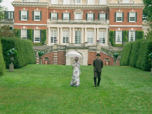 A couple in vintage attire walks on a lawn before a grand mansion. The woman holds a parasol. The setting is lush with green hedges.