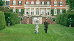 A couple in vintage attire walks on a lawn before a grand mansion. The woman holds a parasol. The setting is lush with green hedges.