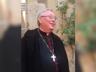 A smiling man in clerical attire with a cross necklace stands outdoors, beige stone wall and greenery in the background.