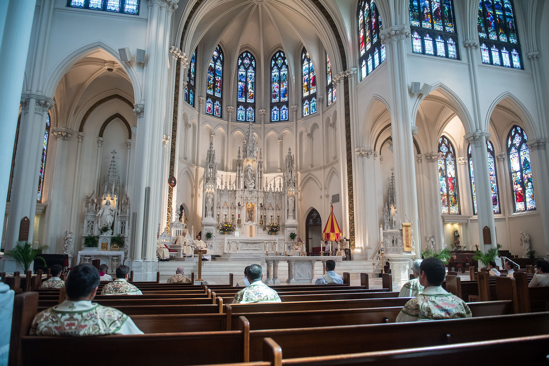 Priests and attendees seated in a grand cathedral with ornate stained glass windows, arched ceilings, and a detailed altar in view.