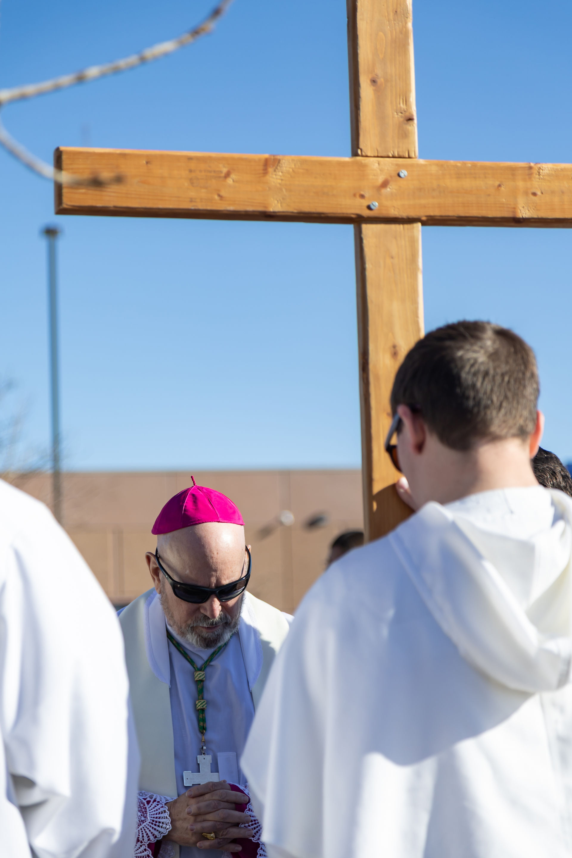 A religious figure in white robes and pink cap prays in front of a large wooden cross. Blue sky and building in the background.