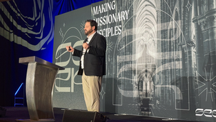 Man speaking on stage with a microphone, in front of a backdrop reading "Making Missionary Disciples." Blue lighting and cathedral image.