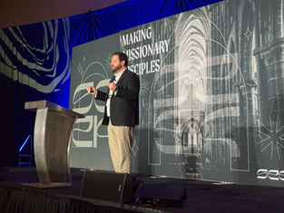 Man speaking on stage with a microphone, in front of a backdrop reading "Making Missionary Disciples." Blue lighting and cathedral image.