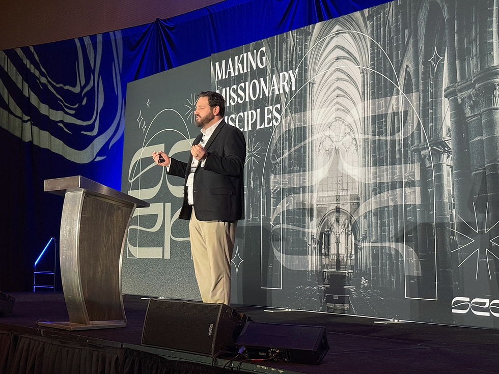 Man speaking on stage with a microphone, in front of a backdrop reading "Making Missionary Disciples." Blue lighting and cathedral image.