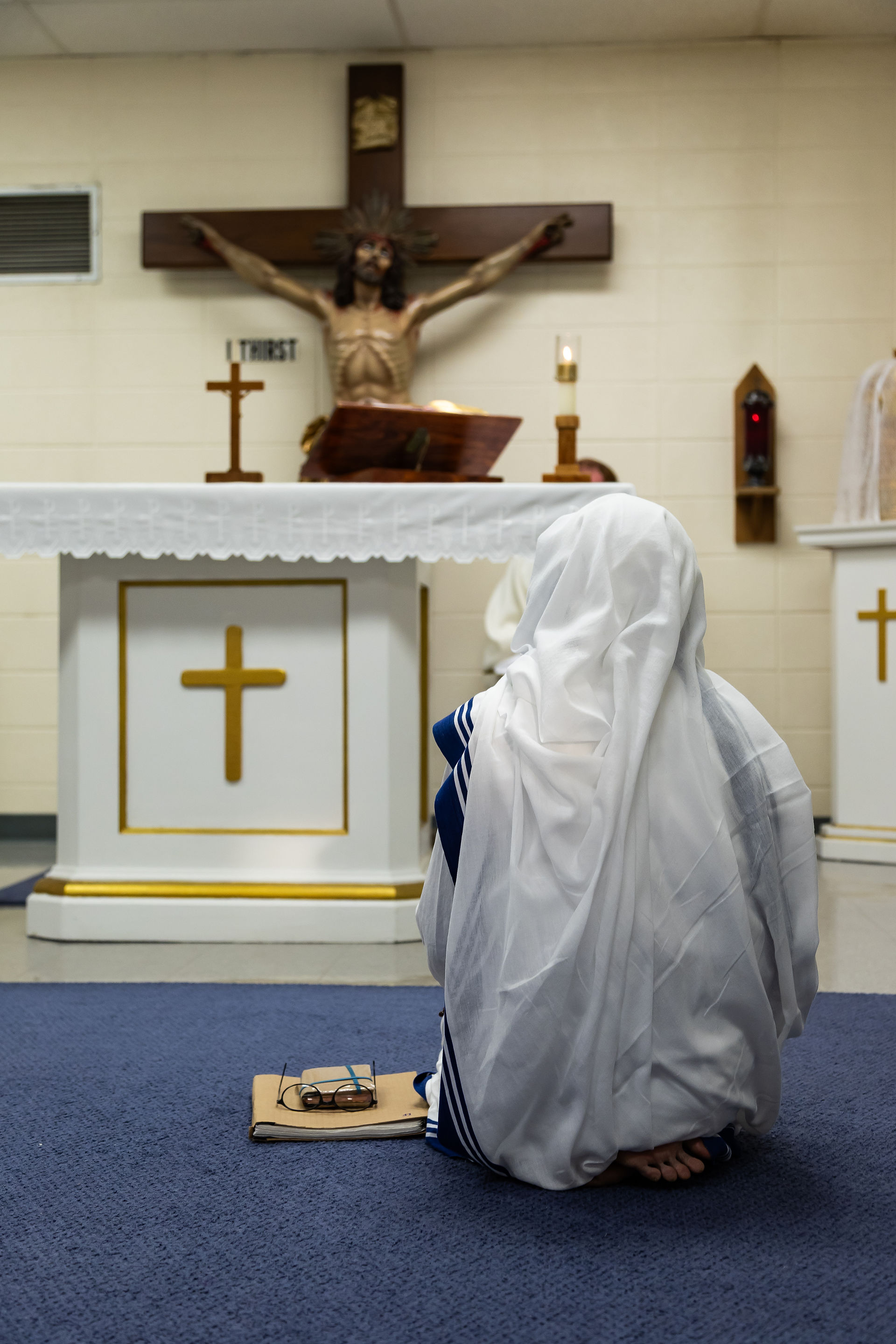 A person in a white habit kneels on a blue carpet, facing an altar with a crucifix. Glasses rest on a book nearby, creating a solemn mood.
