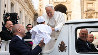 A smiling religious leader in white greets a child in a white outfit, held by a smiling man. They're near an ornate building, filmed by a cameraman.
