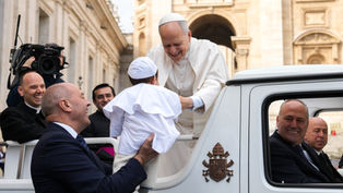 A smiling religious leader in white greets a child in a white outfit, held by a smiling man. They're near an ornate building, filmed by a cameraman.
