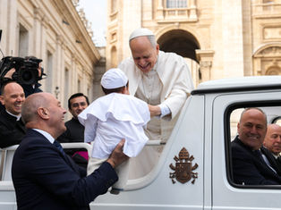 A smiling religious leader in white greets a child in a white outfit, held by a smiling man. They're near an ornate building, filmed by a cameraman.