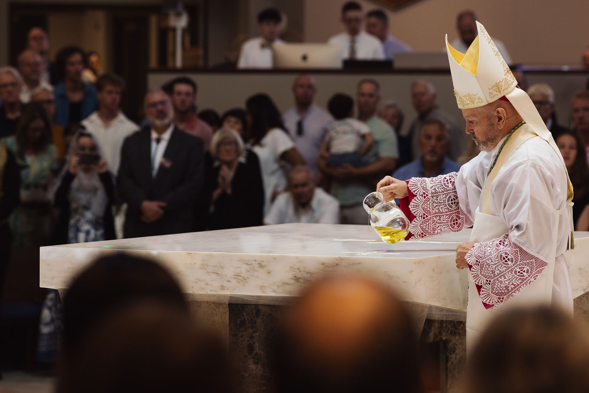 A bishop in ornate robes pours oil on a marble altar during a ceremony, with a congregation watching. The setting is a crowded church.