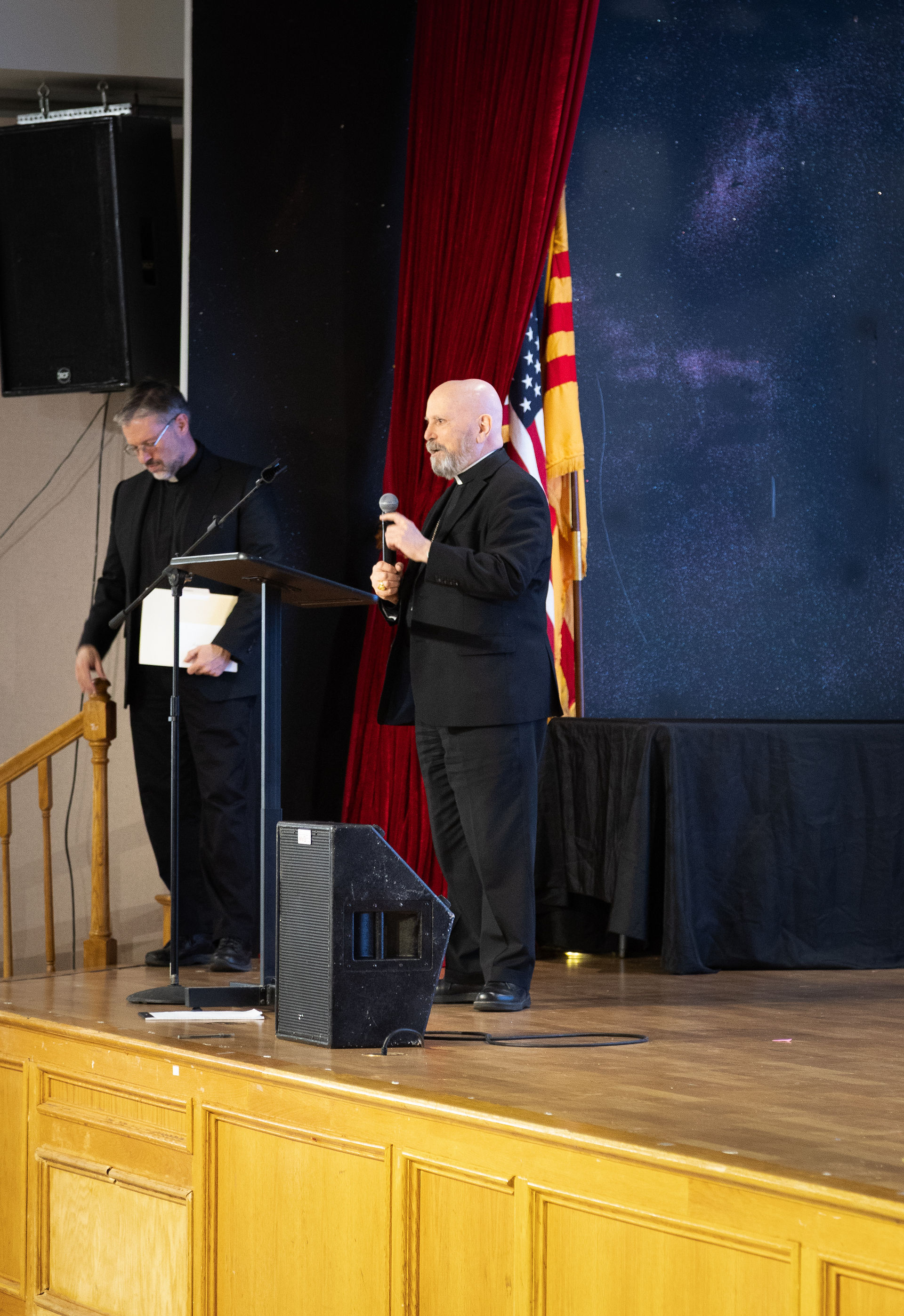 Two men in black suits stand on a stage; one holds a microphone at a podium. Red curtain and flag backdrop, wooden floor, and speaker visible.