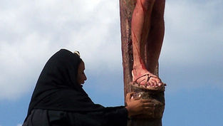 A woman in black kneels at a cross, touching a man's feet, which are bleeding. The sky is cloudy, conveying a solemn mood.