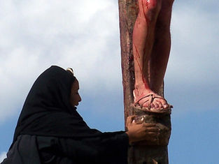 A woman in black kneels at a cross, touching a man's feet, which are bleeding. The sky is cloudy, conveying a solemn mood.
