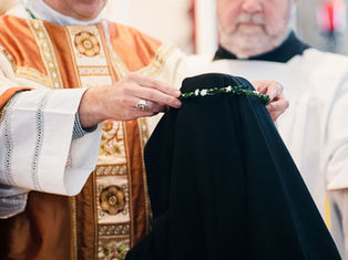 A person in ornate robes places a green floral crown on a figure in black cloth. The setting is solemn and ceremonial.