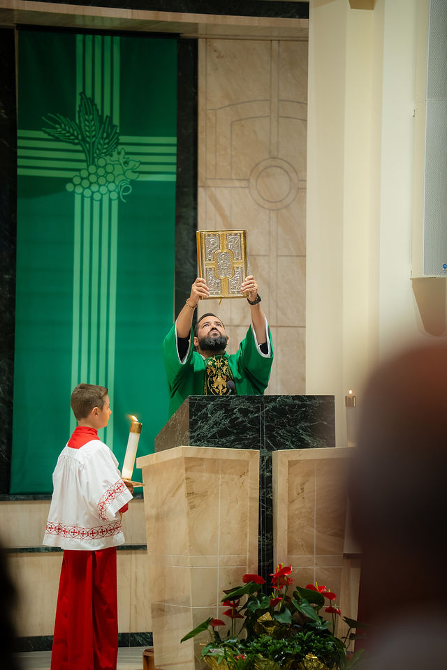 Deacon in green vestments lifts a gold book at the altar while an altar boy stands beside a candle in a solemn church interior.