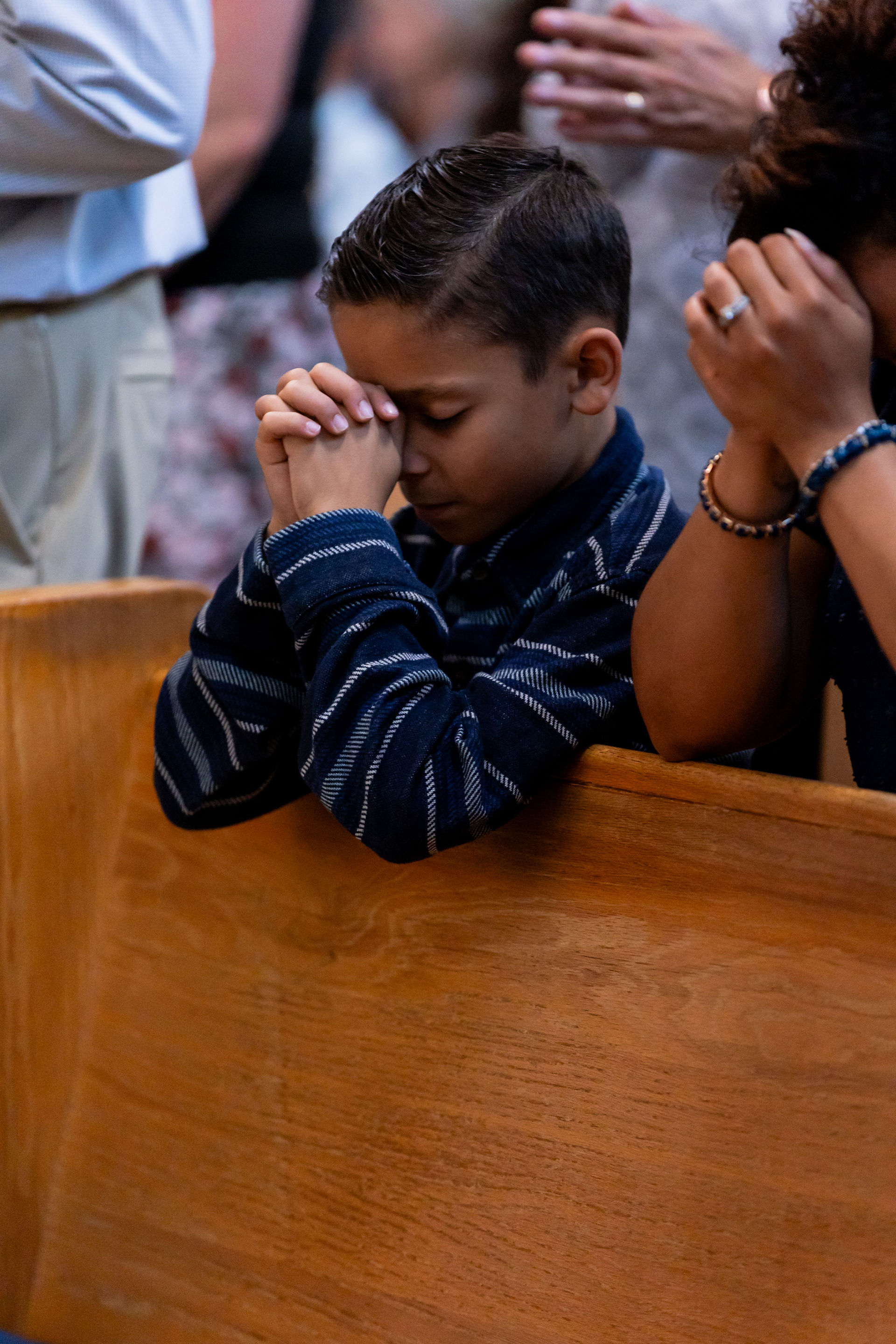 Boy in striped shirt prays with clasped hands in a wooden church pew, surrounded by others with similar gestures. Serene atmosphere.
