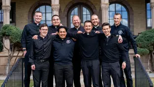 Group of nine smiling men in black clothing stand arm-in-arm outside a building with arches. The mood is joyful and relaxed.