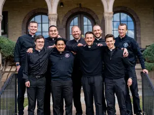 Group of nine smiling men in black clothing stand arm-in-arm outside a building with arches. The mood is joyful and relaxed.