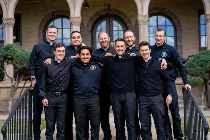Group of nine smiling men in black clothing stand arm-in-arm outside a building with arches. The mood is joyful and relaxed.